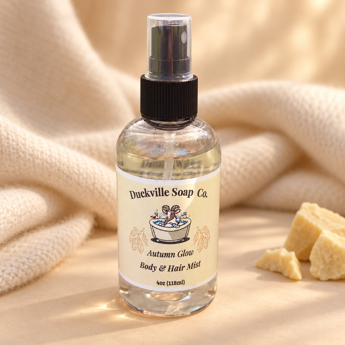 Autumn Glow Duckville Body & Hair Mist bottle on a cream-colored surface with soft fabric and cocoa butter pieces.